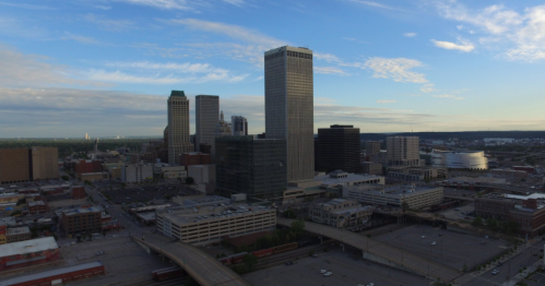 Aerial view of a city skyline featuring tall buildings and a clear blue sky, with roads and parking lots below.