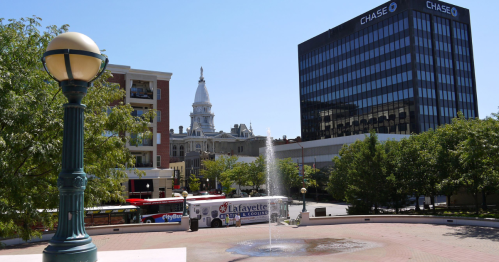 A city square featuring a fountain, buildings, and a bus, with a clear blue sky in the background.