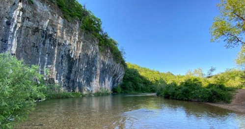 A serene river flows beside a rocky cliff, surrounded by lush greenery under a clear blue sky.