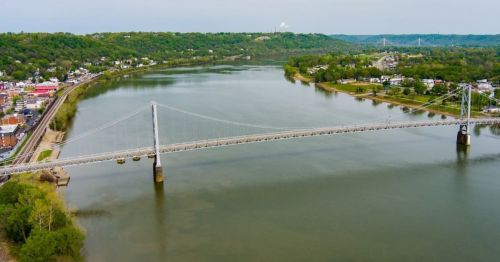 Aerial view of a river with a suspension bridge, surrounded by green hills and a small town along the shore.