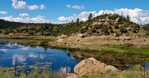 A serene landscape featuring a calm lake, rocky hills, and lush greenery under a blue sky with fluffy clouds.