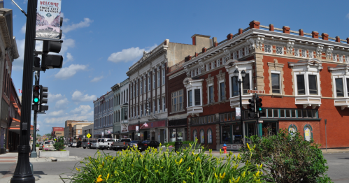 A vibrant downtown street with historic buildings, traffic lights, and greenery under a blue sky with scattered clouds.