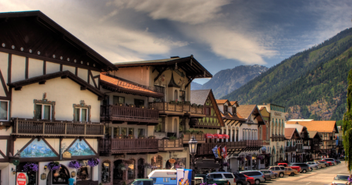 Charming alpine-style buildings line a street with mountains in the background under a blue sky with wispy clouds.