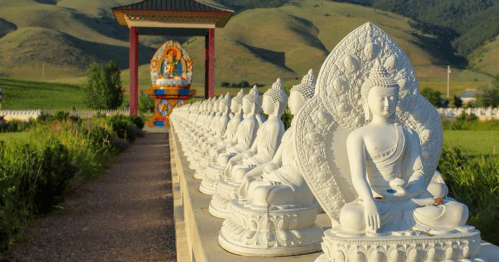 A row of white Buddha statues leads to a colorful temple against a backdrop of green hills.