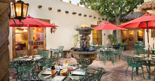 A charming outdoor dining area with green chairs, red umbrellas, and a fountain, surrounded by rustic walls.