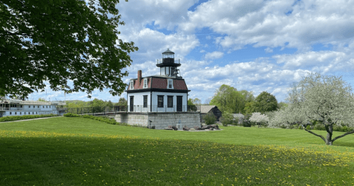 A historic lighthouse on a grassy hill, surrounded by trees and blooming flowers under a partly cloudy sky.
