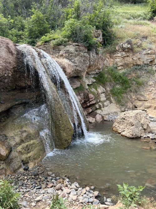 A small waterfall cascades over rocky cliffs into a serene pool, surrounded by lush greenery and natural terrain.