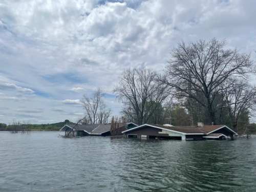 A flooded area with partially submerged houses and bare trees under a cloudy sky.