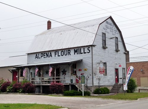 Historic Alpena Flour Mills building with a metal roof, featuring American flags and a sign on a cloudy day.