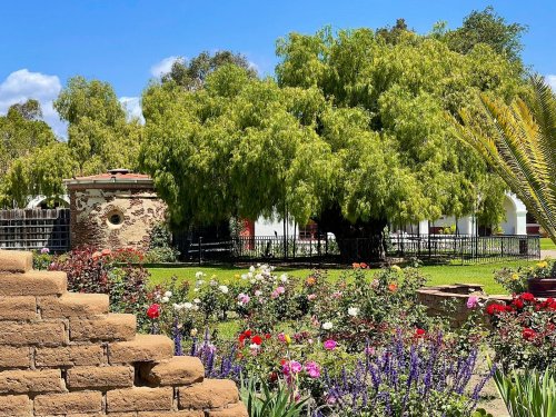 Lush garden with colorful flowers, a large green tree, and a stone structure under a clear blue sky.