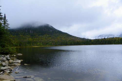 A serene lake surrounded by mountains and trees, with misty clouds hovering above the landscape.