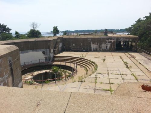 Abandoned concrete structure with circular seating, overgrown with plants, overlooking a body of water in the background.