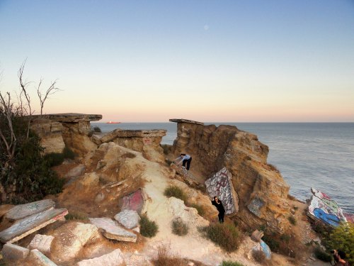 Two people explore rocky cliffs by the ocean at sunset, with graffiti on the rocks and a calm sea in the background.