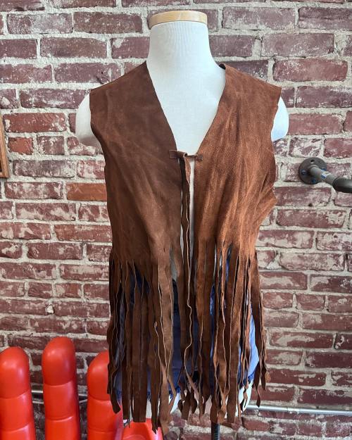 A brown suede vest with long fringes displayed on a mannequin against a brick wall background.