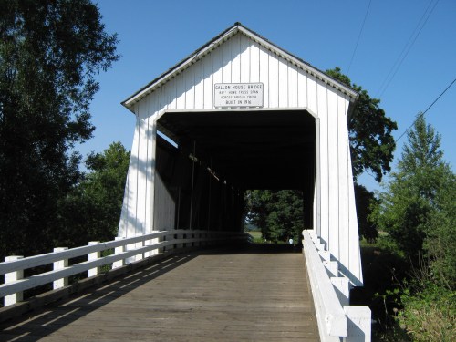 A white covered bridge with a wooden walkway, surrounded by trees and blue sky, labeled "Galloway House Bridge."