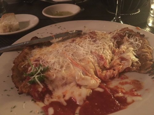 A plate of cheesy chicken parmesan topped with marinara sauce, garnished with parsley, served with bread on the side.