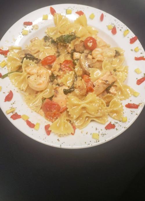 A plate of farfalle pasta with shrimp, cherry tomatoes, spinach, and mushrooms in a creamy sauce, garnished with herbs.