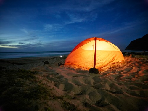 A glowing orange tent on a sandy beach at dusk, with the ocean and a dark cliff in the background.