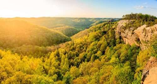 A scenic view of a lush green valley at sunset, with rolling hills and a rocky cliff in the foreground.