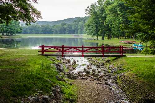 A red wooden bridge spans a rocky stream leading to a calm lake surrounded by lush green trees.