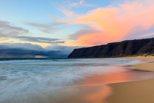 A serene beach at sunset, with gentle waves lapping at the shore and colorful clouds above a distant cliff.