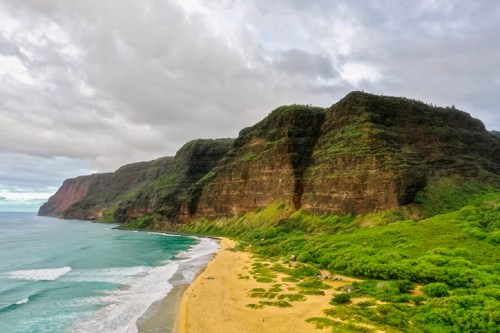 A scenic view of a lush green coastline with cliffs, sandy beach, and gentle waves under a cloudy sky.