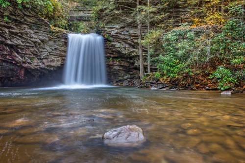 A serene waterfall cascades over rocky cliffs into a calm pool, surrounded by lush greenery and autumn foliage.