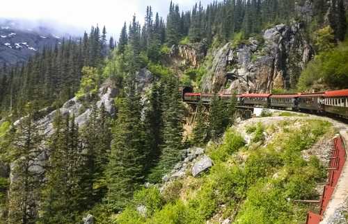 A red train winds through a lush green forest, surrounded by rocky cliffs and mountains under a cloudy sky.