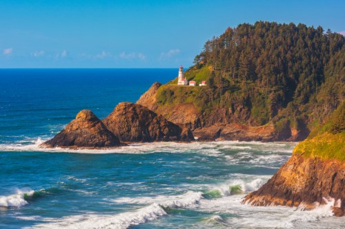 A scenic coastal view featuring a lighthouse on a rocky cliff, surrounded by waves and lush greenery under a clear blue sky.