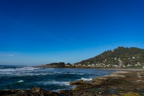 Coastal view with waves crashing on rocks, a clear blue sky, and a hillside dotted with houses in the background.