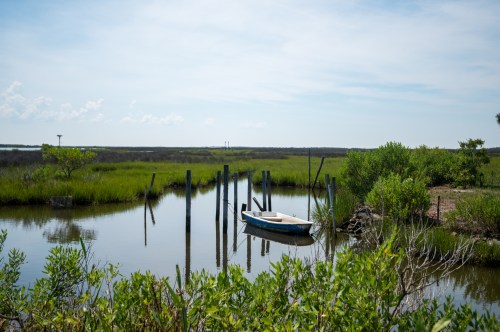 A small boat is moored by wooden posts in a calm, green marshland under a clear blue sky.