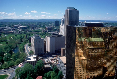 Aerial view of a city skyline featuring modern buildings, green parks, and a river under a clear blue sky.