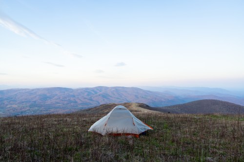 A tent set up on a grassy hilltop, overlooking rolling mountains under a clear sky at dusk.