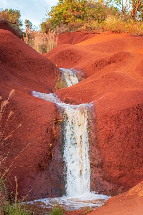 A small waterfall cascades over red, textured earth surrounded by green vegetation under a clear sky.