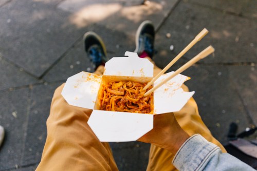 A person holding a takeout box of noodles with chopsticks, sitting on a pavement.