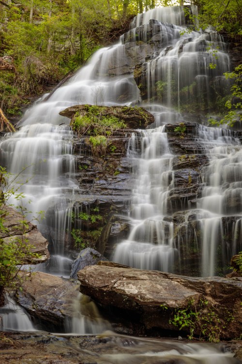 A serene waterfall cascades over rocky ledges, surrounded by lush greenery and soft, flowing water.