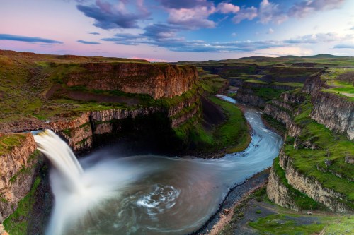 A stunning waterfall cascades into a winding river, surrounded by lush green cliffs under a colorful sky.