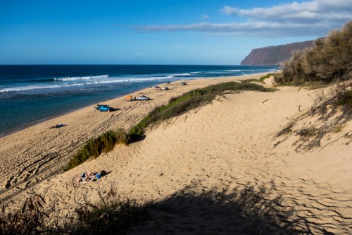 A sandy beach with gentle waves, scattered boats, and a backdrop of cliffs under a clear blue sky.