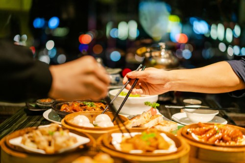 A table filled with various dishes, with hands using chopsticks to pick up food in a vibrant, blurred background.