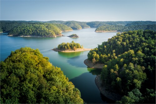 Aerial view of a serene lake surrounded by lush green hills and small islands under a clear blue sky.