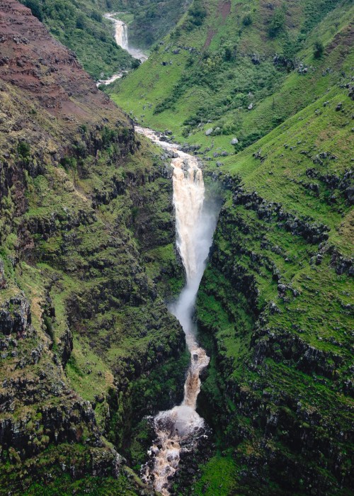 A stunning waterfall cascades down a lush green canyon, surrounded by steep, rocky cliffs and mist rising from the water.