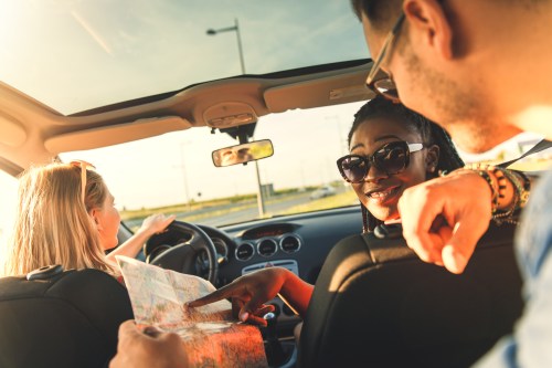 A group of friends in a car, one driving while another looks at a map, enjoying a sunny day.