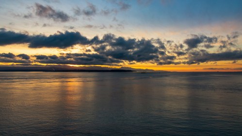 A serene sunset over calm waters, with clouds reflecting warm hues in the sky and a distant shoreline.
