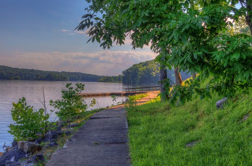 A serene lakeside view with a concrete path, lush greenery, and a wooden dock under a clear sky.