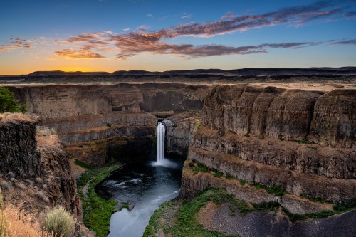 A stunning waterfall cascades into a canyon at sunset, surrounded by rugged cliffs and lush greenery.