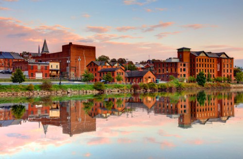 A serene river scene at sunrise, featuring historic brick buildings reflecting on calm water.