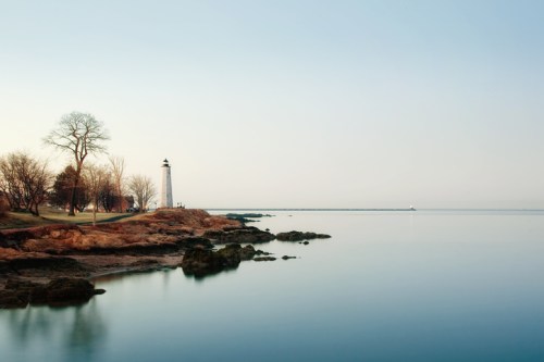 A serene coastal scene featuring a lighthouse, calm waters, and bare trees along the rocky shore at dusk.