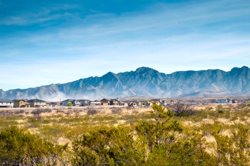 A desert landscape with scattered houses in the foreground and a mountain range in the background under a clear blue sky.