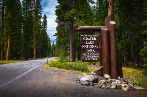 Sign for Crater Lake National Park at the south entrance, surrounded by tall trees and a paved road.