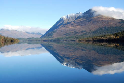 A serene lake reflects snow-capped mountains and a clear blue sky, surrounded by lush greenery.